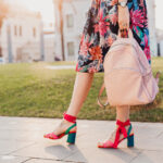 close up details of legs in pink sandals of stylish woman walking in city street in printed colorful skirt, holding pink leather backpack, summer style footwear trend